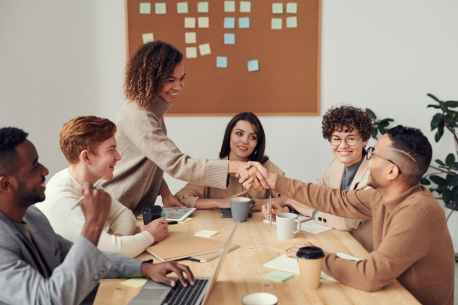 A group meeting around the desk with a female and male shaking hands across the table.